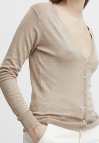 Cardigan beige en tissu tricoté doux, avec un décolleté en V profond, des manches longues et six boutons à l'avant, ainsi que des poignets et un ourlet côtelés.
