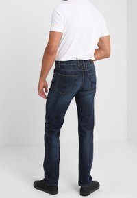 Jeans de denim azul con un corte recto, que presentan bolsillos traseros y un lavado oscuro, combinados con una camiseta blanca y zapatos negros.