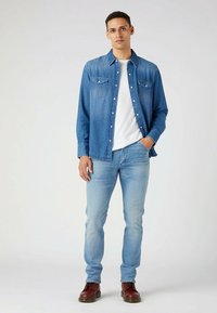 Camisa de denim en azul claro con cuello puntiagudo, dos bolsillos en el pecho y cierres de botón, combinada con jeans azul claro y zapatos marrones.
