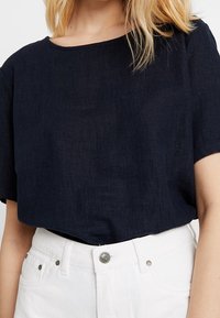 Námořnická modrá lněná halenka s širokým výstřihem a krátkými rukávy, spárovaná s bílými kalhotami s vysokým pasem, které mají detail stříbrného knoflíku.