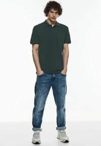 Polo shirt vert foncé à coupe ajustée, à manches courtes et avec col. Porté avec un jean en denim bleu clair et des baskets grises. Design simple.