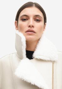 Cappotto in ecopelle bianca con un morbido colletto bianco, caratterizzato da una zip diagonale e dettagli in tessuto testurizzato. I dettagli includono cuciture decorative.
