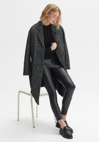 Black and white houndstooth oversized coat with a collar, paired with shiny black leggings and black loafers, worn while seated on a chair.