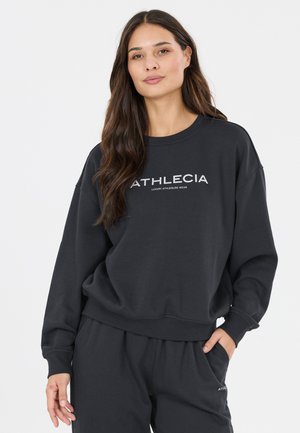 Kvinde med langt mørkt hår iført sort Athlecia sweatshirt og bukser, poserer med den ene hånd i lommen mod hvid baggrund.