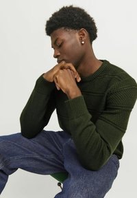 Pull en maille vert foncé avec des motifs texturés et des poignets côtelés, associé à un jean en denim bleu, assis contre un fond uni.