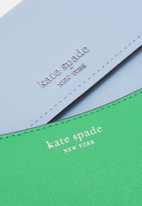 Due portafogli Kate Spade New York, uno blu chiaro con logo argento e uno verde con logo bianco, mostrati da vicino con materiale testurizzato.