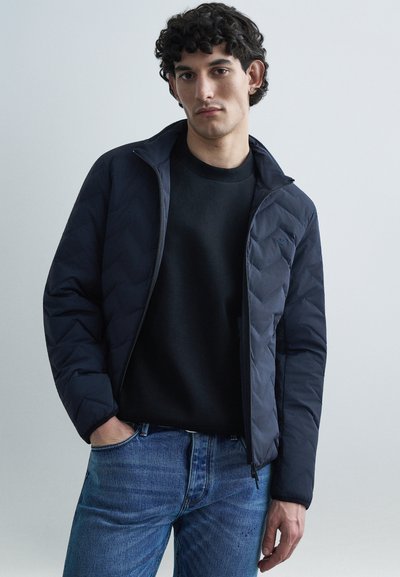Veste matelassée marine avec fermeture éclair et col montant, portée sur un sweat-shirt noir et un jean bleu, présentant un motif en chevrons.