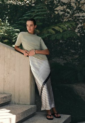 Nő világoszöld rövid ujjú felsőben és fehér pöttyös szoknyában, aki betonos lépcsőkorlátnak támaszkodik, zöld növényzet veszi körül.