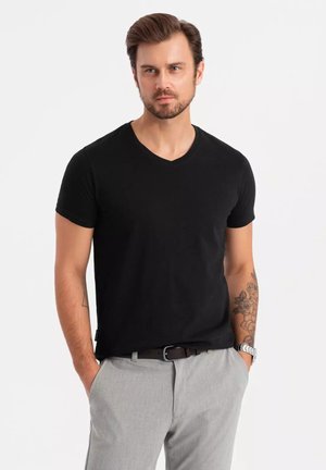 Camiseta negra de cuello en V, hecha de tejido de algodón suave; de manga corta y diseño ajustado. Lleva pantalones gris claro y accesorios minimalistas.