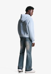 Hellblauer Hoodie mit einem großen Sternmuster auf dem Rücken, kombiniert mit hellen Jeans. Das Model trägt weiße Sneakers. Insgesamt glatte Textur.