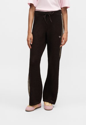 Žena u crnim širokim hlačama s teksturom, bež bočnih pruga i ružičastim tenisicama sa žutim vezicama, stoji na bijeloj podlozi.