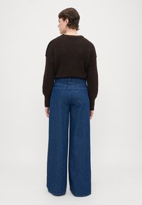 Pulover tricotat maro și blugi denim largi de culoare albastră, prezentând un talie înaltă și buzunare pe spate, afișați din spate pe un fundal deschis.