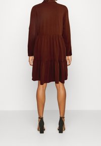 Robe marron à volants en tissu léger, avec manches longues et col arrondi, accompagnée de sandales à talons noirs.