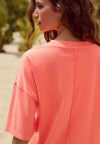 T-shirt oversize color corallo con maniche corte, cucitura testurizzata al centro della schiena, tessuto morbido e vestibilità rilassata. Ambiente esterno visibile.