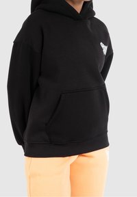 Felpa nera con cappuccio in un tessuto morbido, caratterizzata da una tasca a kangaroo frontale e un logo bianco sul lato sinistro del petto. Abbinata a pantaloni arancioni.