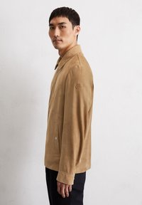 Tan suède jacket met een kraag, zijzakken en een subtiel logo op de mouw. Het ontwerp heeft een gladde textuur en een getailleerde silhouet.