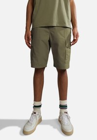 Pantaloni cargo verde oliva con tasche laterali, vestibilità dritta e tessuto leggero, abbinati a scarpe da ginnastica bianche e calzini a righe.