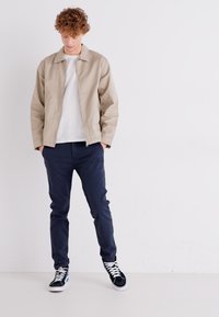 Lys beige lynlåshættejakke over en hvid t-shirt; marineblå slim-fit bukser; sorte sneakers med hvide detaljer; ensfarvet baggrund.