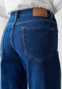 Gros plan sur la poche arrière d'un jean en denim bleu et d'une étiquette de marque en cuir marron, portée avec un haut blanc rentré.