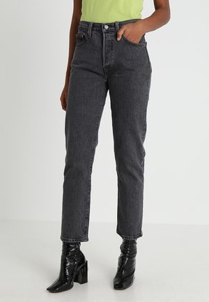 Person trägt eine schwarze High-Waist-Jeans, hat eine Hand in der Tasche und trägt schwarze Stiefeletten mit Absatz, steht auf weißem Boden vor einem weißen Hintergrund.