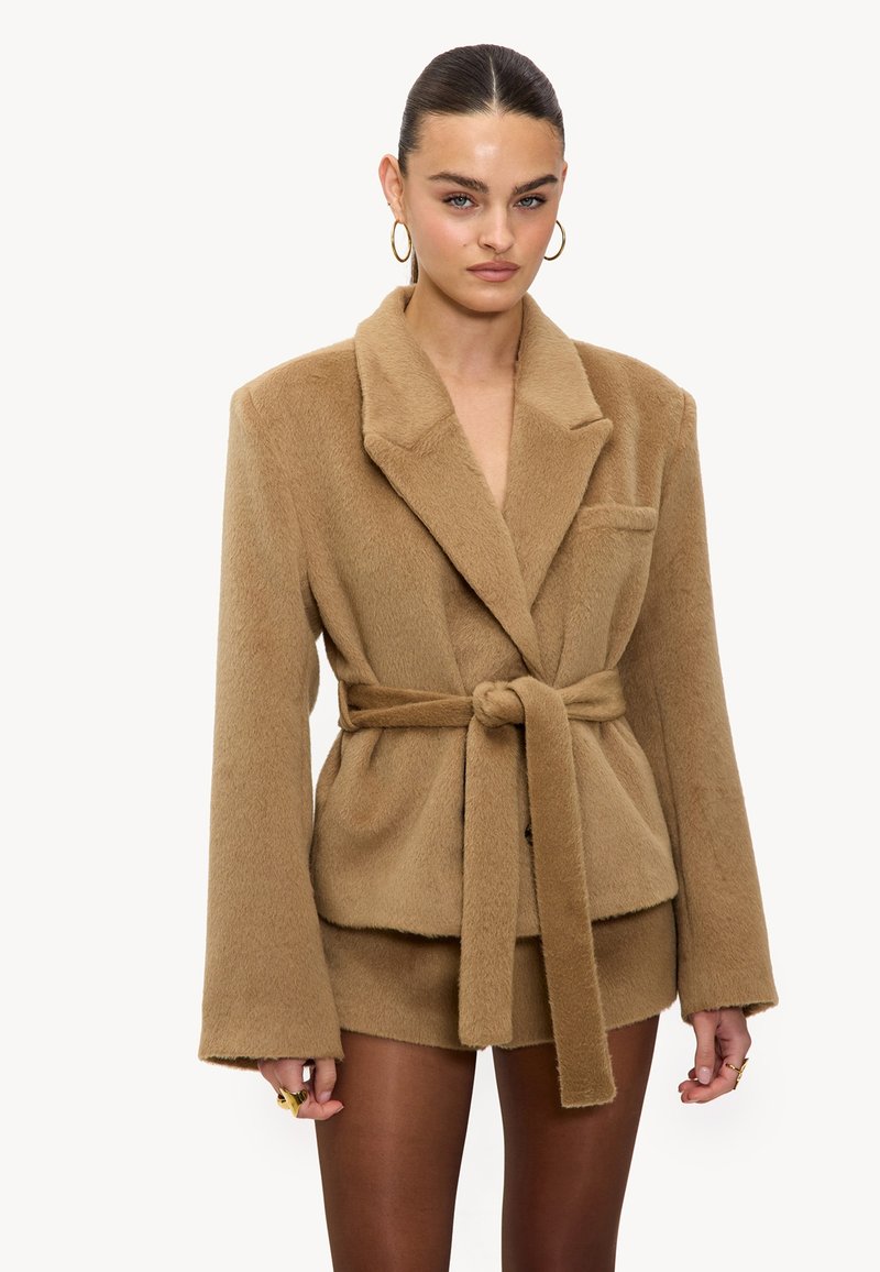 Blazer ceinturé en peluche taupe avec col à revers, manches oversize, poche avant et texture douce. Stylé avec un bas assorti.