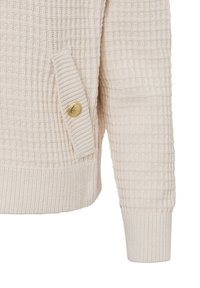 Crème-kleurige, gestructureerde gebreide cardigan met een geribde onderrand, voorzien van een zijzak en een gouden knoopdetail.