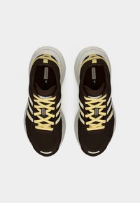 Tumepruunide kollaste paelte ja valge tallaga Adidas Adizero jooksutossude paar, nähtud ülevalt valgel taustal.