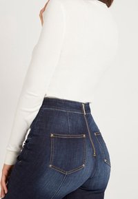 Jean taille haute bleu foncé avec une texture lisse, coutures dorées et une fermeture éclair dissimulée. Comprend des poches arrière avec des coutures courbes.