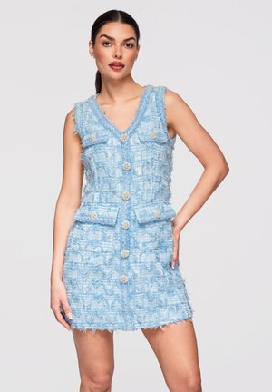 Kobieta w bezrękawowej, jasnoniebieskiej sukience o fakturze, z ozdobnymi guzikami i czterema kieszeniami z patkami, stojąca na jednolitym tle.