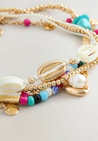 Mehrlagige Armbänder mit goldenen Perlen, weißen Muscheln, mehrfarbigen kleinen Perlen, Türkissteinen und einem Perlenanhänger auf weißen Bändern.