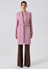 Cappotto tailorato rosa con cintura e rever a punta, abbinato a pantaloni a gamba larga marroni. Tessuto liscio, silhouette strutturata e dettagli minimi.