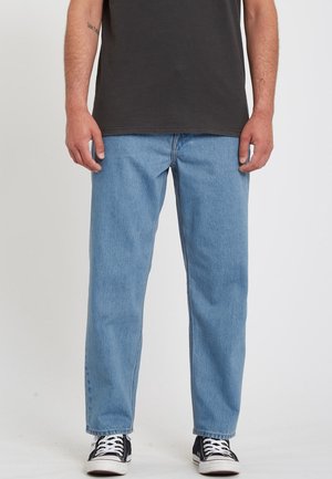 Mężczyzna w jasno-niebieskich dżinsach o prostych nogawkach, czarnym T-shircie oraz czarno-białych trampkach, stojący na jednolitym jasnym tle.