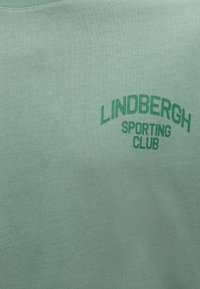 Πράσινο κείμενο "LINDBERGH SPORTING CLUB" τυπωμένο σε ανοιχτό πράσινο ύφασμα.