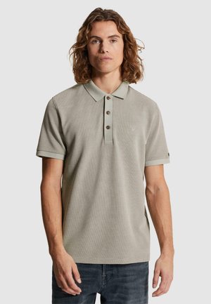 Mann mit schulterlangem lockigem Haar, der ein hellgraues gemustertes kurzärmeliges Polohemd und dunkle Jeans trägt, vor einem einfarbigen Hintergrund stehend.