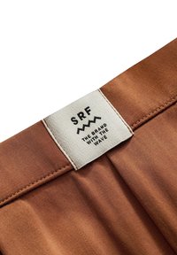 Brązkowa etykieta z materiału z logo "SRF" i napisem "MARKA Z FALĄ". Gładka faktura, obszyte krawędzie i subtelny połysk.