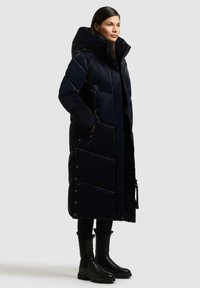 Marineblå pufferjakke med oversized hætte, quiltet design, sidelommer og trykknapdetaljer, parret med sorte ankelstøvler og bukser.