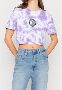 Ustřižené tričko s barevným vzorem v levandulové barvě, s kulatým logem obsahujícím text "TOMMY JEANS SINCE 1985" v zelené a černé barvě. Krátké rukávy a jemná textura.