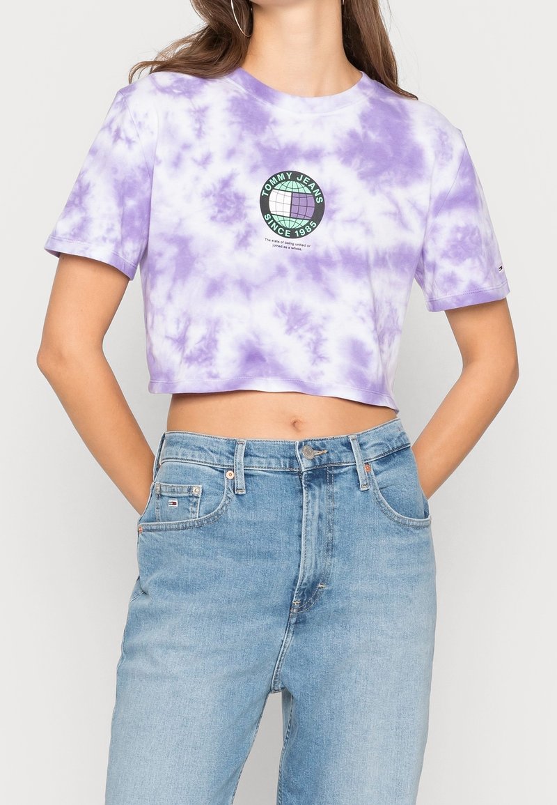 Ustřižené tričko s barevným vzorem v levandulové barvě, s kulatým logem obsahujícím text "TOMMY JEANS SINCE 1985" v zelené a černé barvě. Krátké rukávy a jemná textura.