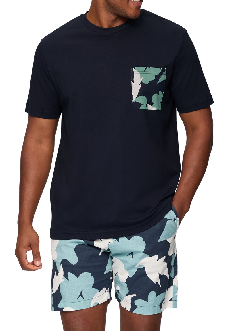 Marineblauw katoenen T-shirt met een bloemenpatroon op de zak, gecombineerd met lichtblauwe shorts met een opvallend bloemenontwerp in wit en marineblauwe accenten.