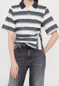 Personne portant un polo à manches courtes bleu marine et blanc à rayures horizontales, rentré dans un jean noir délavé taille haute.