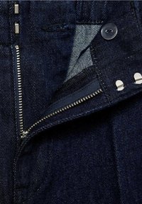 Jean en denim bleu foncé avec fermeture éclair argentée partiellement ouverte, bouton noir, et deux crochets argentés à la ceinture.