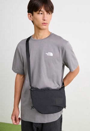 Jeune homme portant un t-shirt gris The North Face et un sac bandoulière noir, regardant vers sa gauche sur un fond uni.