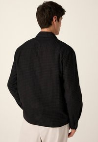 Schwarzes, strukturiertes Hemd mit lässigem Schnitt, Button-Down-Kragen und langen Ärmeln. Verfügt über ein dezentes geometrisches Muster und Knopfmanschetten.