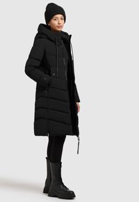 Giacca a vento nera con cappuccio, caratterizzata da un design trapuntato e lacci regolabili. Indossata sopra leggings neri e stivali alti fino al ginocchio con lacci.