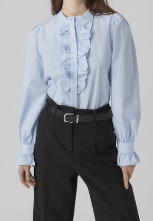 Femme portant un chemisier bleu clair à volants avec des manches bouffantes, rentré dans un pantalon noir taille haute avec une ceinture noire.
