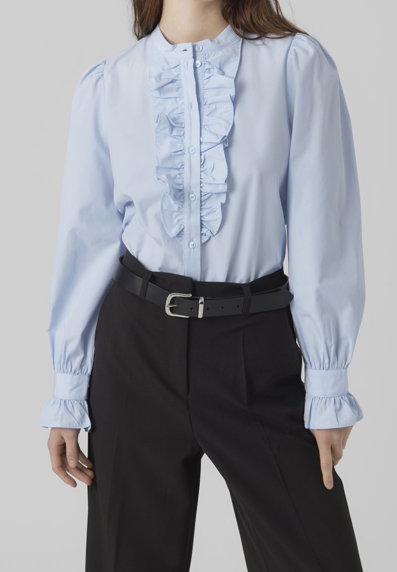 Femme portant un chemisier bleu clair à volants avec des manches bouffantes, rentré dans un pantalon noir taille haute avec une ceinture noire.