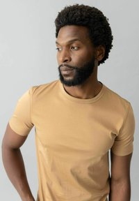 Vīrietis ar cirtainiem matiem un bārdu, tērpies bēšā T-kreklā, skatās uz sāniem uz pelēka fona.