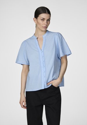 Blusa azzurra chiara a maniche corte con colletto alla cinese, chiusura a bottoni e tessuto texturizzato con motivi sottili. Abbinata a pantaloni neri.