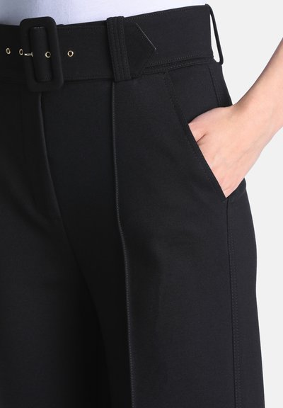 Main portant un pantalon noir avec une ceinture noire assortie, doigts placés dans la poche avant droite.