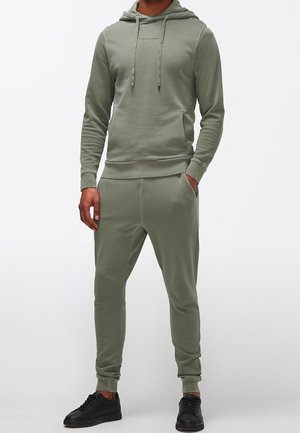 Set di felpa e pantaloni tuta verde oliva in tessuto morbido e elasticizzato. Caratterizzato da cordini, tasche laterali e pantaloni a gamba affusolata. Sneakers nere completano il look.
